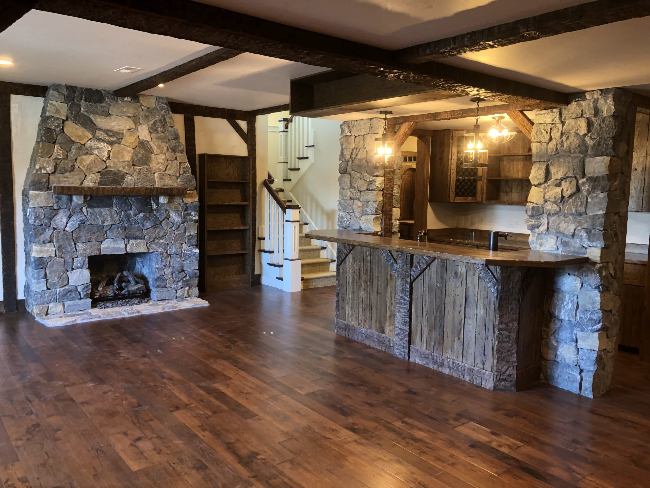 Rustic basement pub with stone fireplace, hand-hewn beams, and reclaimed-wood bar — Harlan Custom Contracting project